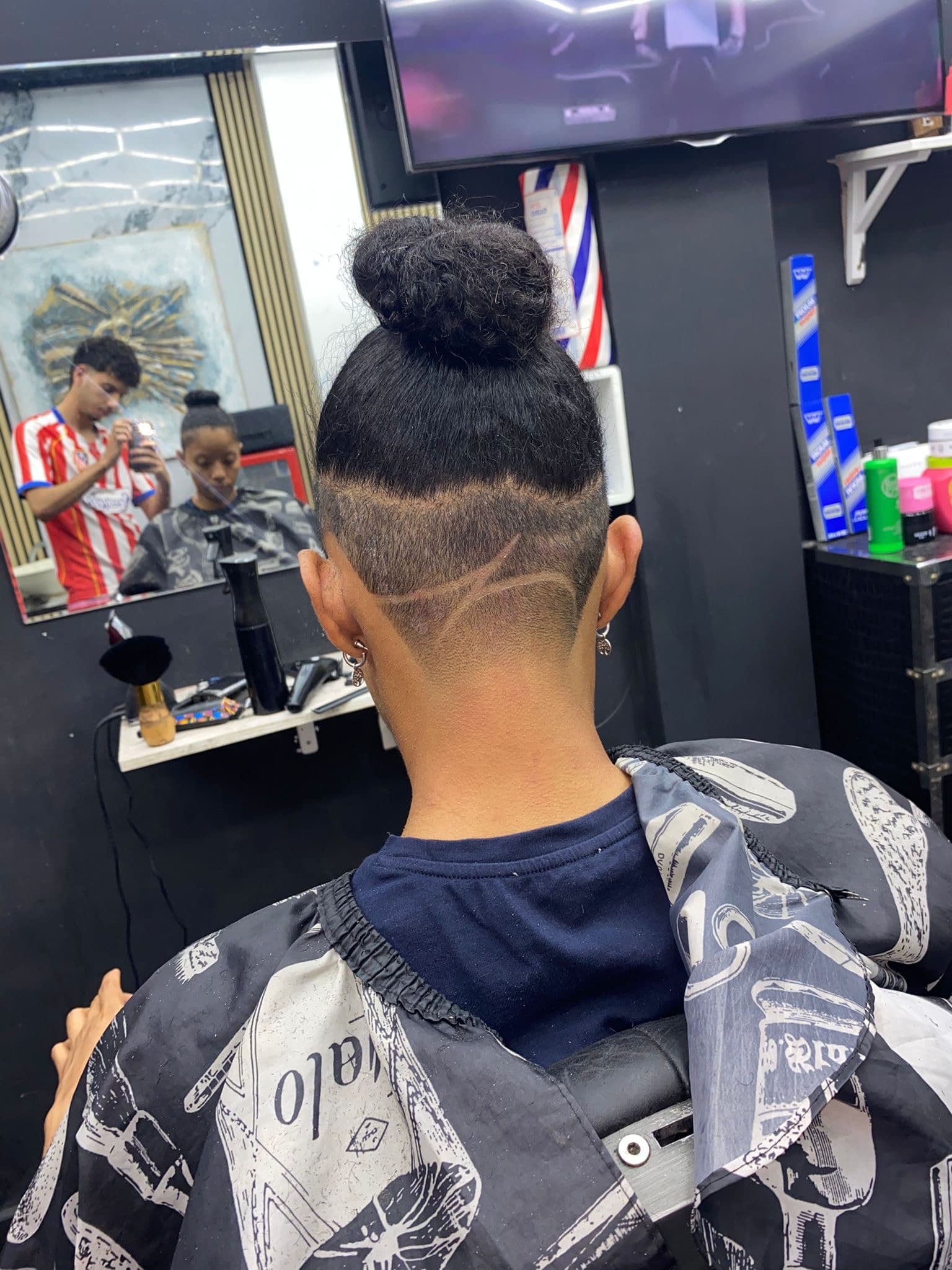 Moño alto y rapado inferior con diseño de líneas curvas en una barbería, vista trasera.